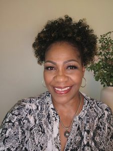 Smiling professional woman with curly hair, wearing hoop earrings and a patterned blouse, posing in front of a light-colored wall with a decorative plant in the background.